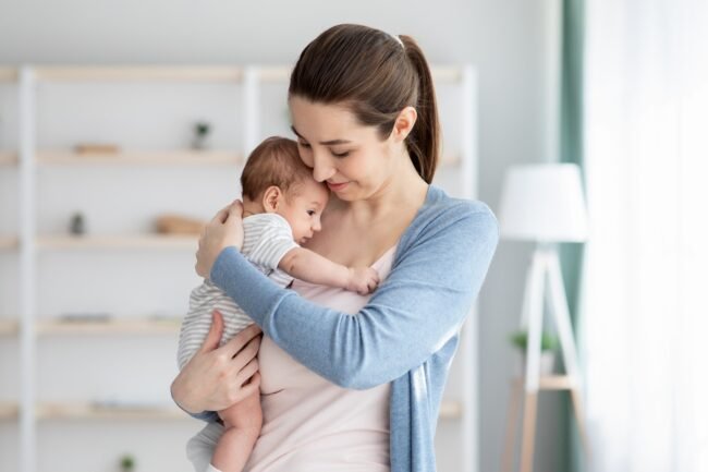 Young Smiling Mum Holding Her Adorable Newborn Baby, Tenderly Embracing Her Child